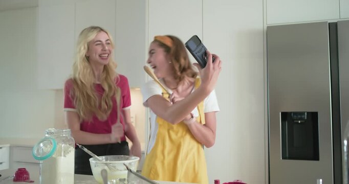 Female friends in kitchen taking selfie checking cake recipe on phone and mixing batter with whisk