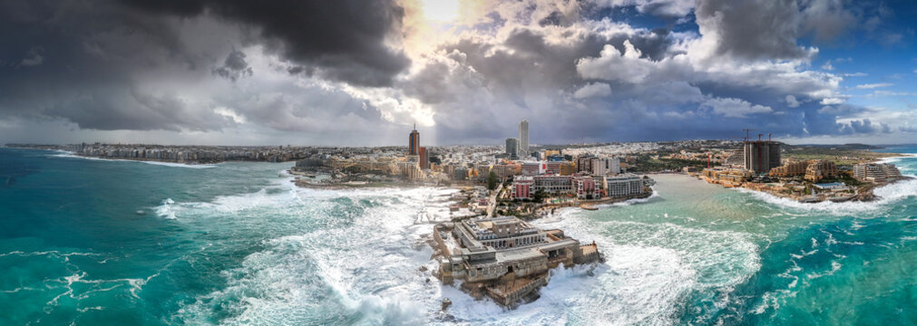 Aerial view of the dramatic cityscape where the sea crashes against the fortress walls, contrasting with the skyline under a stormy sky, St. Julian's, Malta.