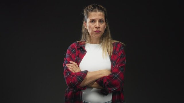 Woman with arms crossed wearing red plaid flannel shirt and white tee in studio, visible forearms and folded pose against black backdrop; disapproval.