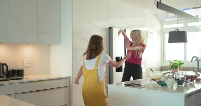 Female friends picking up wooden spoons and dancing around central island in kitchen as microphones