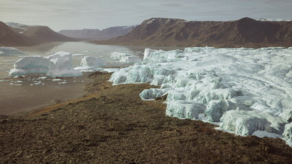 Glistening ice formations rise majestically from the calm waters of a glacial lake. Mountains loom in the distance, casting shadows on rocky terrain under soft morning light. © icetray