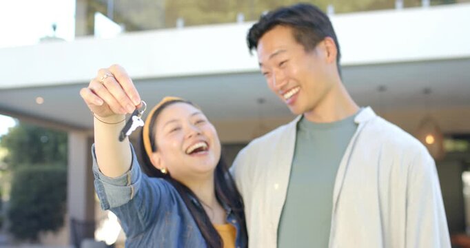 Asian couple celebrating new home, woman wearing denim holding keyring at porch, man embracing her