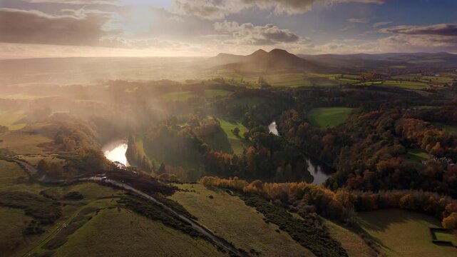 Aerial view of the twisting River Tweed, flanked by autumnal trees, leading towards the Eildon Hills under a sunlit sky, Melrose, Scotland, United Kingdom.