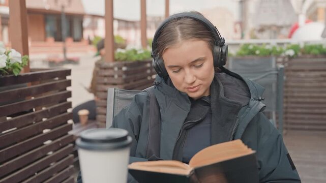 Caucasian woman reading outdoors wearing headphones, sipping coffee at urban cafe terrace, flipping paperback pages while bundled in dark jacket, thoughtful expression, closeup facial shots with soft