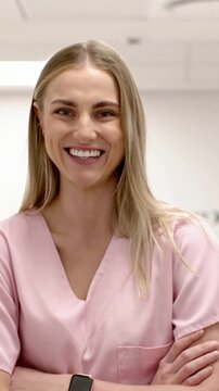 Vertical video: Smiling woman in pink scrubs smartwatch arms crossed clinic reacting to camera zoom