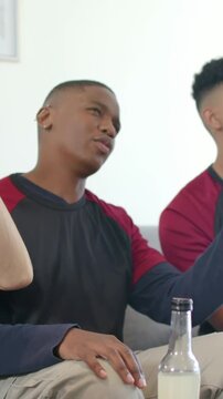 Vertical video: Gesturing man reacting to friend on couch at home, with bottle and raglan shirt