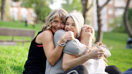 Three happy senior women friends laughing and embracing while sitting on green grass in a public park, enjoying togetherness