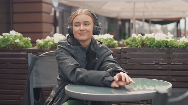 young white woman at outdoor cafe wearing black jacket and headphones, seated at round table beside planter boxes, smiling and thoughtful while listening to music or podcast frames show student,