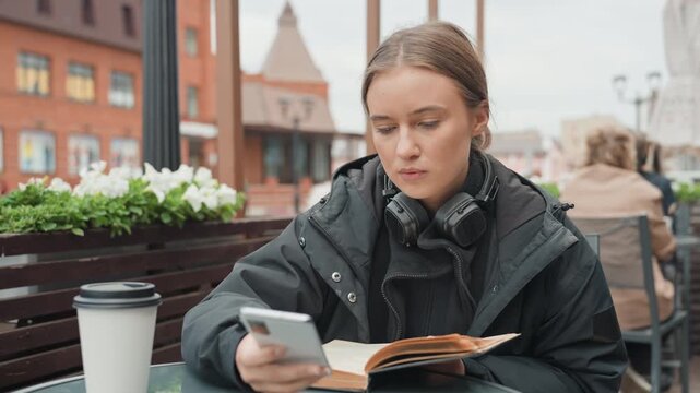 Caucasian woman reading notebook outdoors cafe, studious freelancer alternating between writing and phone calls, headset resting around neck, warm jacket and scarf, takeaway coffee cup on table,
