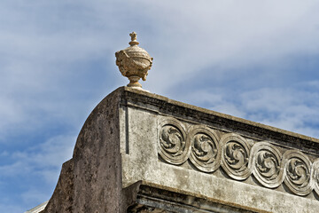 Obraz premium detail in Leirosa, Portugal, showcasing classical stonework with a sculpted urn or finial crowning the upper edge. Beneath it, a frieze features a repeating spiral or volute motif, carved into weather