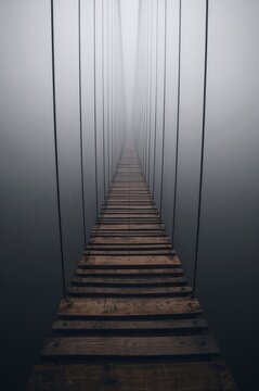 Foggy suspension bridge rises overhead as rusted cables vanish into deep mysterious abyss