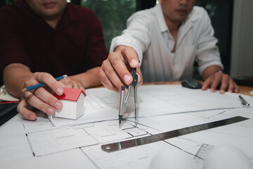 Close up of architect hand using compass divider on construction blueprint. Engineer team designing home structure with house model and ruler.
