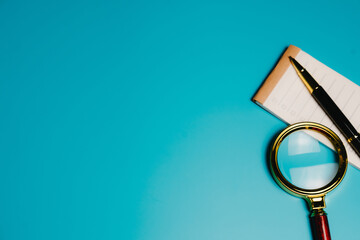 Research concept. Magnifying glass pen notepad blue background office tools close up writing instrument paper investigation study focus detail inspection business