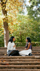 Two indian women friends sitting on park stairs, enjoying autumn outdoors, talking and sharing moments together
