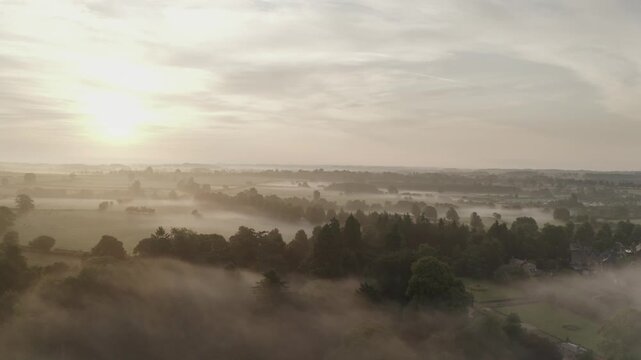 Aerial view of the sun breaking over the horizon to reveal trees and fields covered in a blanket of fog, Ripley, England, United Kingdom.
