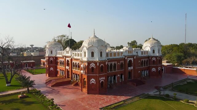 Aerial view of the Noor Mahal palace with its red brick facade contrasting with white domes, surrounded by green lawns and pathways, Bahawalpur, Punjab, Pakistan.