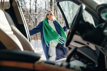 Woman wearing sunglasses and a green scarf stands beside an open car door in a snowy forest, showcasing winter fashion and outdoor adventure