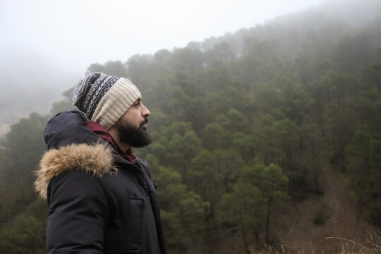 A bearded man contemplates the landscape of trees and mist on a foggy winter day.
