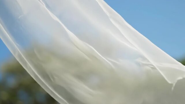 Sheer white fabric gently blows in the wind against a soft blue sky. Lush green trees blur in the background, creating a serene atmosphere.
