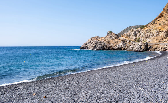 Chios island, Greece. Mavra Volia Beach, black pebble coast. Gentle waves below rugged cliffs, blue sky, sunny day.