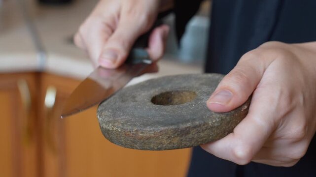 Close-up view of a person's hands carefully sharpening a metal kitchen knife blade using a traditional circular whetstone, restoring its cutting edge for meal preparation