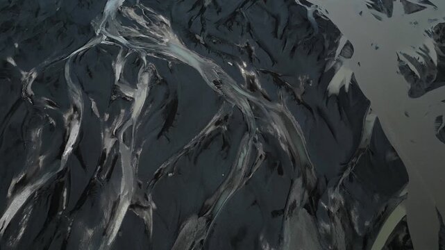 Aerial view of braided glacial river patterns weaving through dark, volcanic terrain, showcasing the stark beauty of Iceland's landscape, Svinafell, Iceland.
