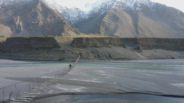 Aerial view of a person crossing the thrilling Hussaini Suspension Bridge over a wide river, framed by towering mountains, Hunza, Gilgit, Pakistan.