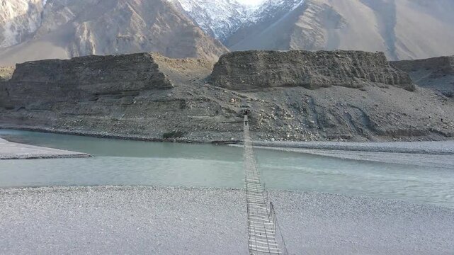 Aerial view of Hussaini Suspension Bridge with people crossing over the grey river surrounded by rocky mountains, Hunza, Gilgit, Pakistan.
