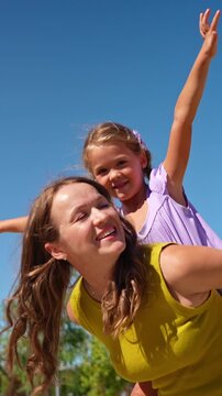 Joyful little girl having fun with her mother on a sunny day. Young woman carrying her cute child on her back against a clear blue sky