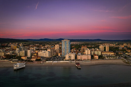 Aerial view of the city skyline meeting the tranquil sea under a vibrant sunset sky, with a prominent skyscraper rising above the urban landscape, Follonica, Tuscany, Italy.