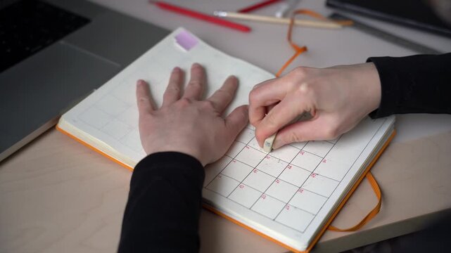 Close-up of female hands with an eraser erasing a date in a clear empty handmade planner calendar, the woman erases dates in an orange planner lying on a table