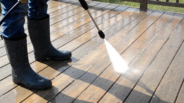 Man in blue jeans and black rubber boots pressure washing dirty wooden deck planks, professional cleaning service
