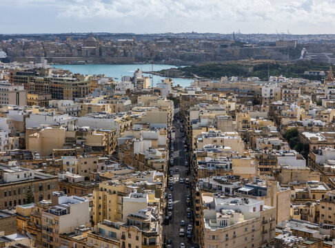 Aerial view of a dense urban landscape with sandstone buildings bordering a narrow street leading toward the azure sea, Valletta, Malta.
