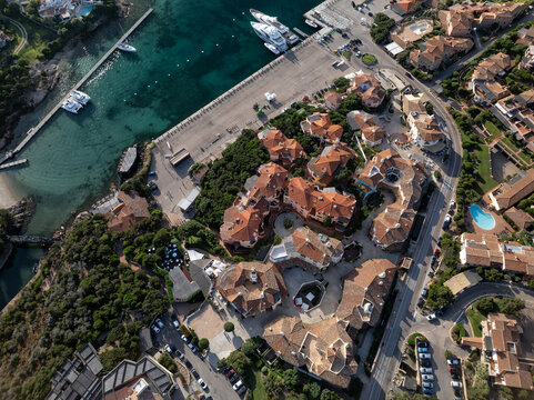Aerial view of terracotta rooftops cascade towards the azure harbor where luxury yachts rest, framed by verdant hills, Porto Cervo, Sardinia, Italy.