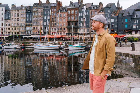 In the historic harbor of Honfleur, a man in a flat cap and yellow shirt gazes across the Vieux Bassin, where traditional sailboats and tall, slate-fronted houses reflect beautifully in the calm water