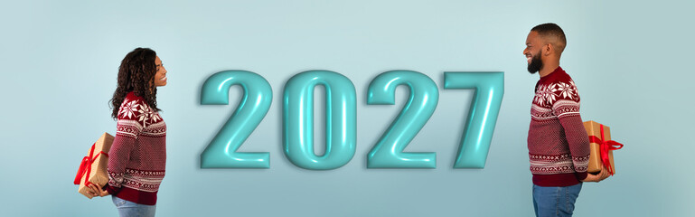 Obraz premium A couple stands facing each other, holding wrapped gifts. They wear festive sweaters and look happy. A large number 2027 is displayed behind them, indicating a celebration.