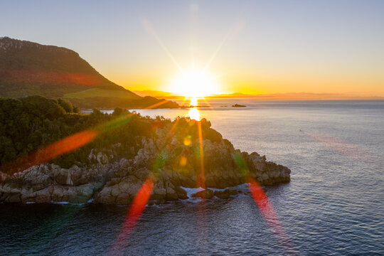 Aerial view of Leisure Island's rocky coastline ablaze with the sun's golden fire, contrasting with the deep blue sea, Mount Maunganui, Bay of Plenty Region, New Zealand.