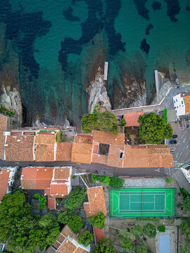 Aerial view of red-tiled roofs meet the turquoise sea, a tennis court nestled among the buildings, Cadaques, Spain.