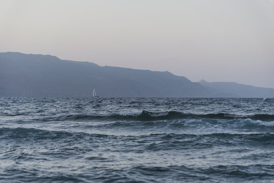 Lonely sailboat sailing on rough sea against misty mountains at twilight