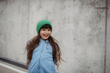 Naklejka premium Outdoor portrait of cute young girl with beanie hat. Girl with long hair standing on playground in the city.