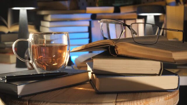 Open book with eyeglasses and tea cup on wooden table indoors, Quiet reading scene featuring literature stack illuminated by warm desk lamps