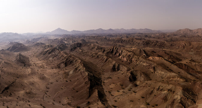 Aerial view of the dramatic, arid landscape, where rugged mountains meet the vast desert under a hazy sky, Wadi Bani Khalid, Ash Sharqiyah North Governorate, Oman.