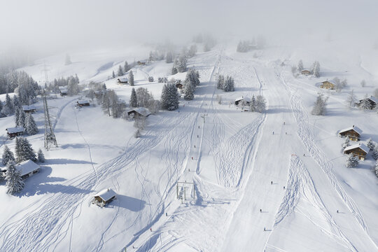 Aerial view of a snowy ski slope winding through Les Mosses, dotted with chalets and shrouded in mist, creating a serene winter wonderland, Ormont-Dessous, Vaud, Switzerland.