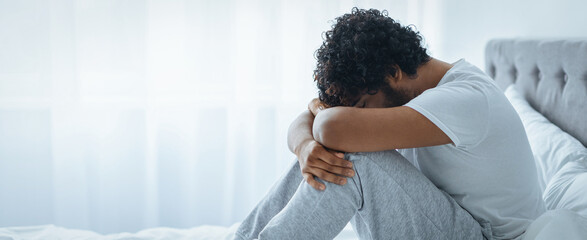 A man sits on the edge of a bed with his head resting on his knees. He appears deep in thought in a...