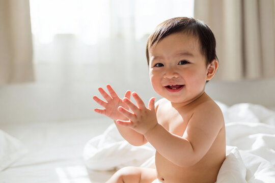 Happy baby with dark hair claps hands joyfully while sitting on white bed in bright room. Natural light creates warm atmosphere, concept of childhood happiness and early development.