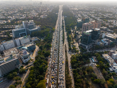 Aerial view of heavy traffic congestion on Delhi - Jaipur Expressway, framed by lush greenery and modern buildings, Gurugram, Haryana, India.