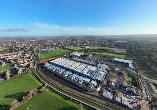 Aerial view of the modern Google AI data centre, a structure of pale blue and white contrasts sharply against the surrounding green fields, Waltham Cross, United Kingdom.