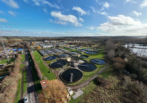 Aerial view of circular sewage tanks glisten under a vast sky, contrasting with the surrounding greenery, Hoddesdon, United Kingdom.