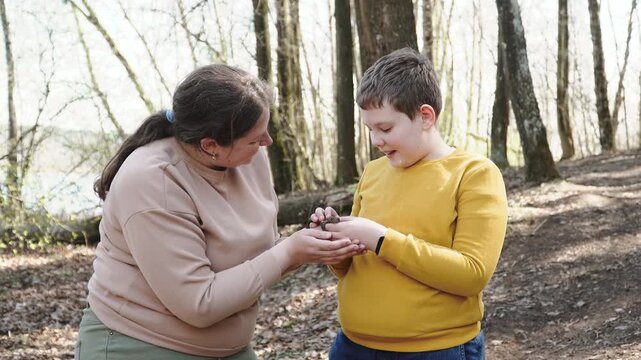 Caucasian woman and boy smiling and holding frog in forest. Studying nature and caring for animals. Portrait of woman and child helping toad during walk on sunny day.