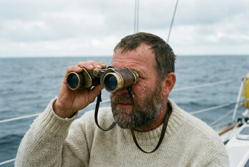 A rugged, bearded sailor in a wool sweater scans the ocean horizon with vintage binoculars from a sailboat. A concept of navigation and adventure.